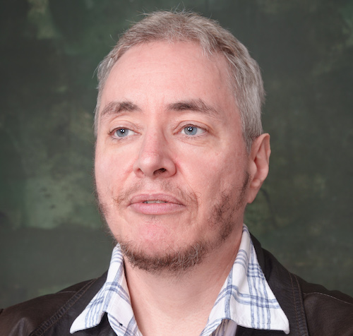 A picture of the author, Kell Shaw, looking into the middle distance. He's in his late forties, with grey hair, a scruffy beard, and cool leather jacket.